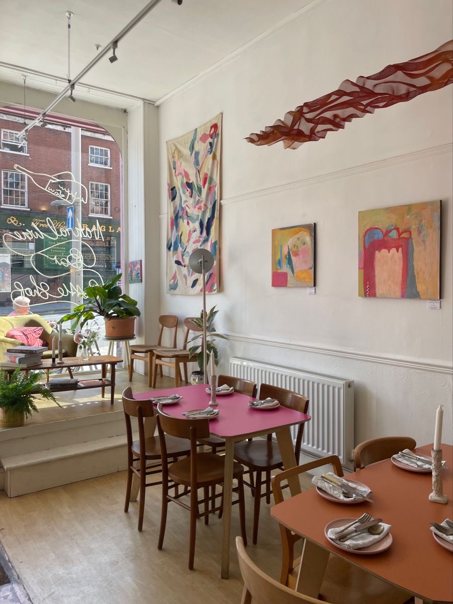 Helles Café-Interieur mit bunten abstrakten Bildern, rosa und terrakotta Tischen, Holzstühlen und Pflanzen