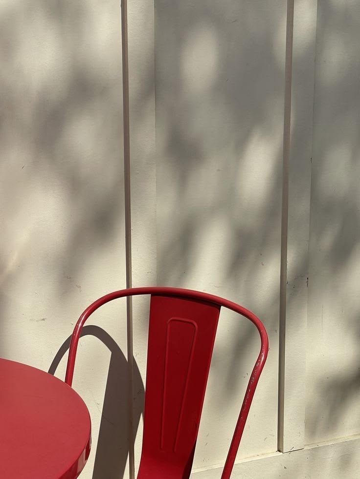 Rotes Metallstuhl und Tisch vor einer hellen Holzwand mit zarten Blattschatten im Sonnenlicht