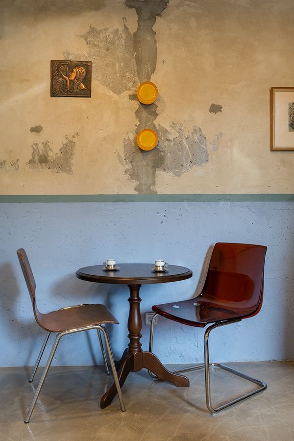 Runder Café-Tisch mit zwei Stühlen vor einer patinierten Wand mit gelben Tellern und Kunst